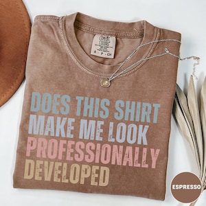 Puede incluir: Una camiseta marr&oacute;n de manga corta con cuello redondo y etiqueta Comfort Colors. La prenda presenta el texto "DOES THIS SHIRT MAKE ME LOOK PROFESSIONALLY DEVELOPED" en azul claro, rosa y beige. Un collar plateado fino descansa sobre la tela.