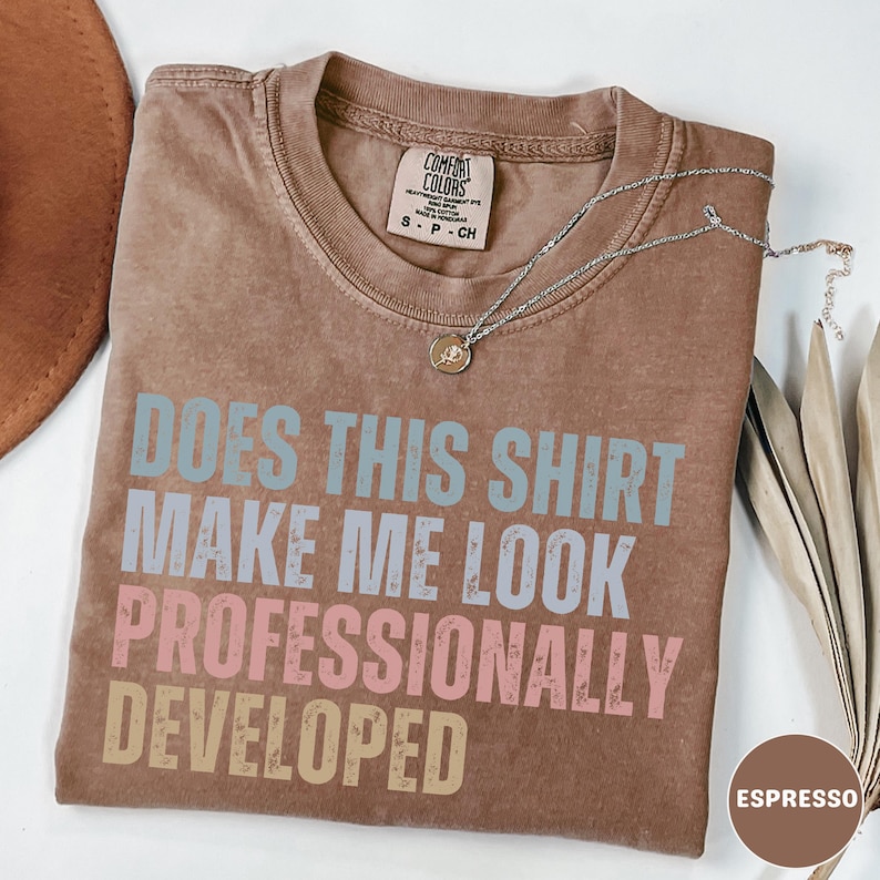 Puede incluir: Una camiseta marr&oacute;n de manga corta con cuello redondo y etiqueta Comfort Colors. La prenda presenta el texto "DOES THIS SHIRT MAKE ME LOOK PROFESSIONALLY DEVELOPED" en azul claro, rosa y beige. Un collar plateado fino descansa sobre la tela.