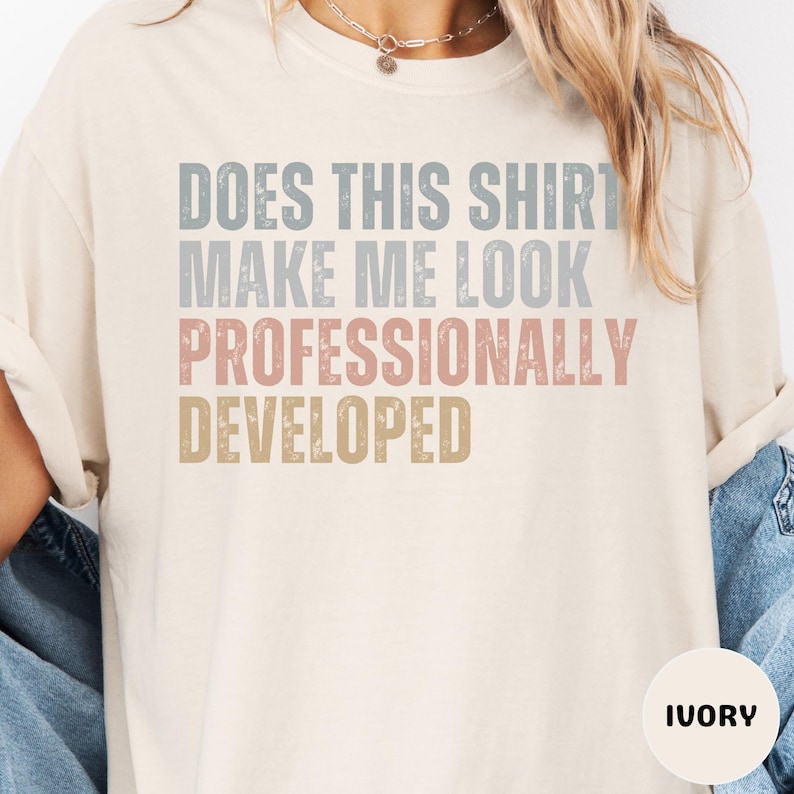 Puede incluir: Una camiseta de manga corta color marfil con el texto desgastado DOES THIS SHIRT MAKE ME LOOK PROFESSIONALLY DEVELOPED en l&iacute;neas apiladas de color azul, gris, rosa y dorado. La camiseta tiene cuello redondo y un corte holgado.