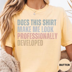 Puede incluir: Una camiseta de manga corta color amarillo mantequilla con el texto desgastado "DOES THIS SHIRT MAKE ME LOOK PROFESSIONALLY DEVELOPED" en letras de bloque azules, rosas y beige. La camiseta tiene cuello redondo y corte holgado.