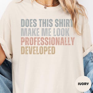 May include: An ivory short-sleeve t-shirt featuring the distressed text DOES THIS SHIRT MAKE ME LOOK PROFESSIONALLY DEVELOPED in stacked lines of blue, grey, pink, and gold. The shirt has a crew neckline and a relaxed fit.