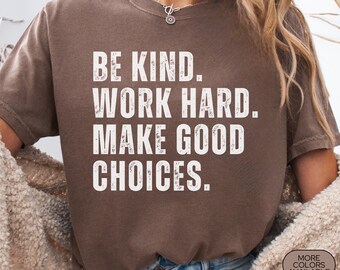 May include: Espresso-colored t-shirt with the text "BE KIND. WORK HARD. MAKE GOOD CHOICES." in white block letters. The shirt is a Comfort Colors brand and has a vintage look. Additional text includes "MORE COLORS AVAILABLE."