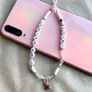 Peut inclure: Une breloque de téléphone perlée blanche avec les mots "ISAIAH 60" et "SHINE" en lettres dorées. Le charme comprend un pendentif en forme d'étoile dorée et repose sur un téléphone rose.
