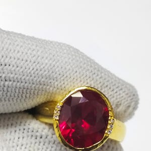 Peut inclure: Une bague en or ornée d'une grosse pierre précieuse rouge foncé de taille ovale. La bague a un anneau en or et est ornée de petites pierres précieuses claires de chaque côté de la pierre principale. Fond blanc.