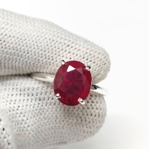 May include: A silver ring with a large, oval-cut ruby gemstone. The ruby is a deep red colour and is held by four prongs. The ring is displayed against a white background.