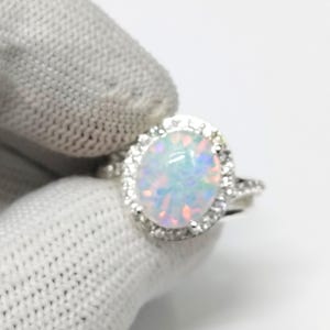 Könnte beinhalten: Ein silberner Ring mit einem großen, ovalen Opal-Edelstein, umgeben von kleinen, klaren Steinen. Der Opal zeigt schillernde Farben von Blau, Rosa und Weiß. Der Ring wird von einer behandschuhten Hand vor weißem Hintergrund gehalten.