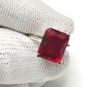 Può includere: Un anello d'argento con una grande gemma rettangolare di colore rosso intenso. La gemma è incastonata con quattro griffe e la fascia dell'anello è sottile e lucida. L'anello è mostrato su sfondo bianco.