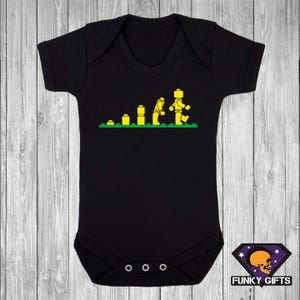 May include: Black baby bodysuit featuring a yellow graphic of a Lego figure's evolution. The design shows the figure assembling from blocks. The bodysuit has short sleeves and three poppers at the bottom. The Funky Gifts logo is in the bottom right corner.