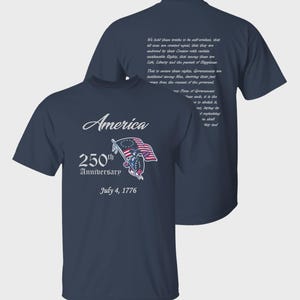 May include: A navy blue short-sleeve t-shirt featuring a graphic on the front with the text America, 250th Anniversary, and July 4, 1776, alongside an illustration of a flag-bearing figure. The back displays a block of white printed text.