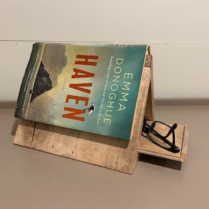 May include: A wooden book stand holding a book titled "Haven" by Emma Donoghue. The book has a teal and orange cover. Black eyeglasses rest on a small shelf below the book. The stand is made of light-coloured wood.