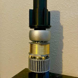 May include: A close-up of a black, silver, and gold lightsaber hilt. The hilt is made up of several different parts, including a black handle, a silver ring, and a gold coil.