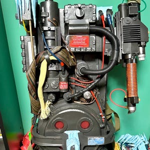 Può includere: Una replica dettagliata di un pacco protonico di Ghostbusters. Il pacco è principalmente grigio scuro con tubi neri, accenti rossi e una cravatta a righe marroni e gialle. Presenta vari componenti meccanici e una bacchetta.