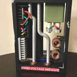 May include: A black box with a clear plastic front, showing a circuit board with wires and components. The box has a red label that reads "HIGH VOLTAGE PRESENT".
