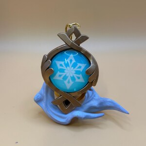 May include: A detailed pendant featuring a blue orb with a white snowflake design, encased in a bronze-colored frame. The pendant is attached to a gold-colored chain and rests on a light blue sculpted base. The overall design is intricate and decorative.