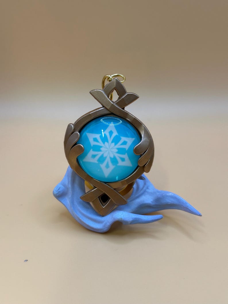 May include: A detailed pendant featuring a blue orb with a white snowflake design, encased in a bronze-colored frame. The pendant is attached to a gold-colored chain and rests on a light blue sculpted base. The overall design is intricate and decorative.