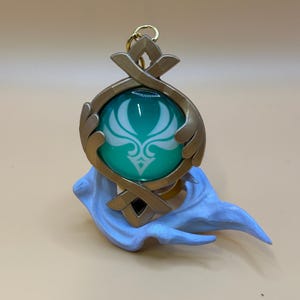 May include: A teal and gold pendant with a white emblem, suspended from a gold-colored ring. The pendant is set on a light blue sculpted base. The design features a teal orb with a white floral pattern, framed by gold-colored metal.