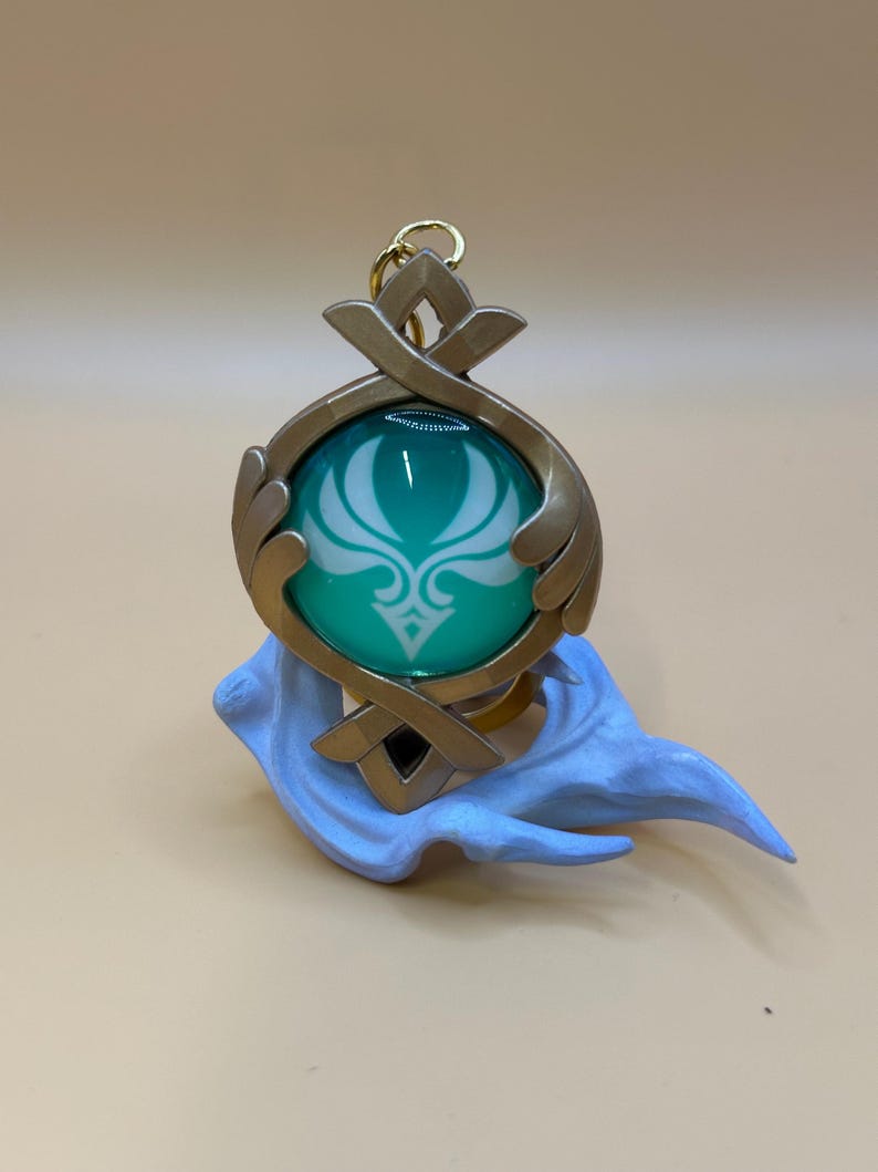 May include: A teal and gold pendant with a white emblem, suspended from a gold-colored ring. The pendant is set on a light blue sculpted base. The design features a teal orb with a white floral pattern, framed by gold-colored metal.
