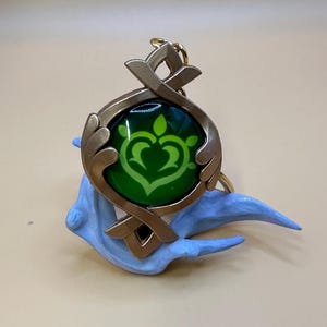 May include: A gold-colored pendant with a green center featuring a stylized heart-shaped design. The pendant is attached to a gold-colored keyring and rests on a light blue sculpted base. The design is reminiscent of a video game.
