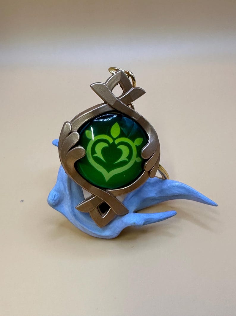 May include: A gold-colored pendant with a green center featuring a stylized heart-shaped design. The pendant is attached to a gold-colored keyring and rests on a light blue sculpted base. The design is reminiscent of a video game.