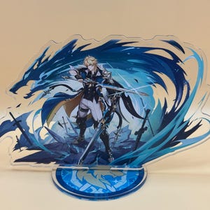 May include: An acrylic standee featuring a blonde character in a white and blue outfit holding a sword, surrounded by swirling blue energy effects and a dragon silhouette. The figure stands on a circular blue base with a printed emblem design.