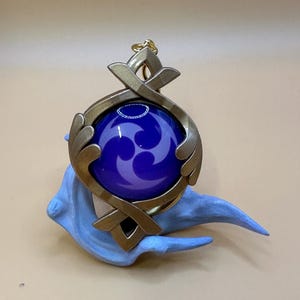 May include: A gold-colored pendant with a dark blue orb featuring a purple swirl design. The pendant is set on a light blue, stylized base. The pendant has a decorative, geometric design.