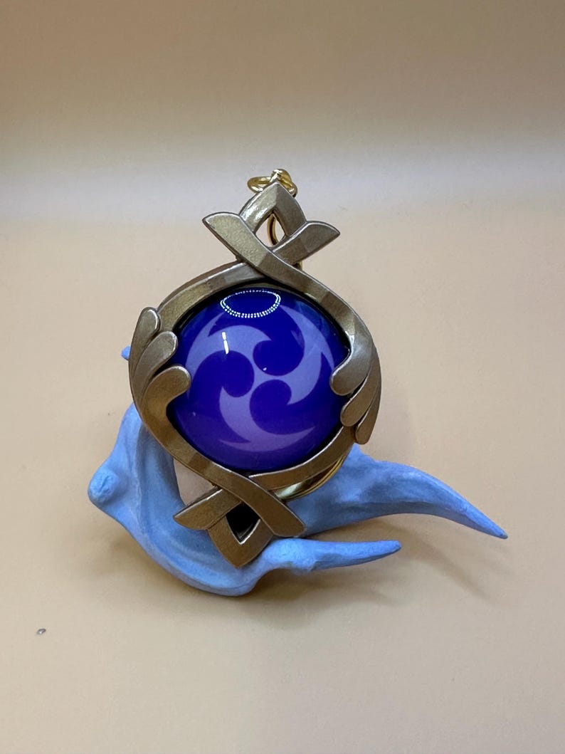 May include: A gold-colored pendant with a dark blue orb featuring a purple swirl design. The pendant is set on a light blue, stylized base. The pendant has a decorative, geometric design.