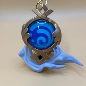 May include: A gold-colored pendant with a blue orb featuring a white swirl design. The pendant has a decorative frame and is attached to a gold-colored loop. A light blue, cloud-like base supports the pendant.
