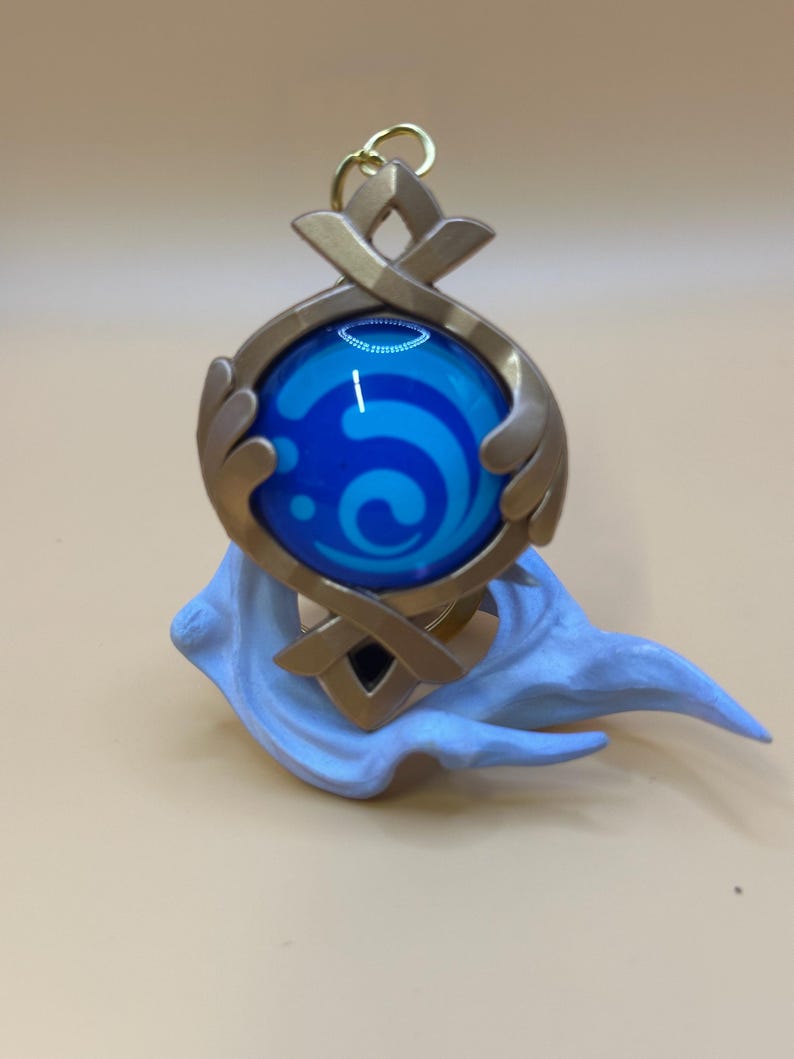 May include: A gold-colored pendant with a blue orb featuring a white swirl design. The pendant has a decorative frame and is attached to a gold-colored loop. A light blue, cloud-like base supports the pendant.