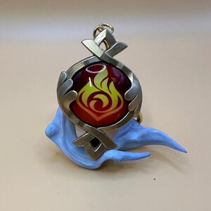May include: A pendant with a fiery design. The pendant features a circular, metallic frame with a red and yellow flame emblem. It is set against a light blue, sculpted base, and includes a gold-colored loop for attachment.
