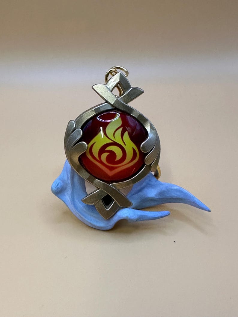 May include: A pendant with a fiery design. The pendant features a circular, metallic frame with a red and yellow flame emblem. It is set against a light blue, sculpted base, and includes a gold-colored loop for attachment.
