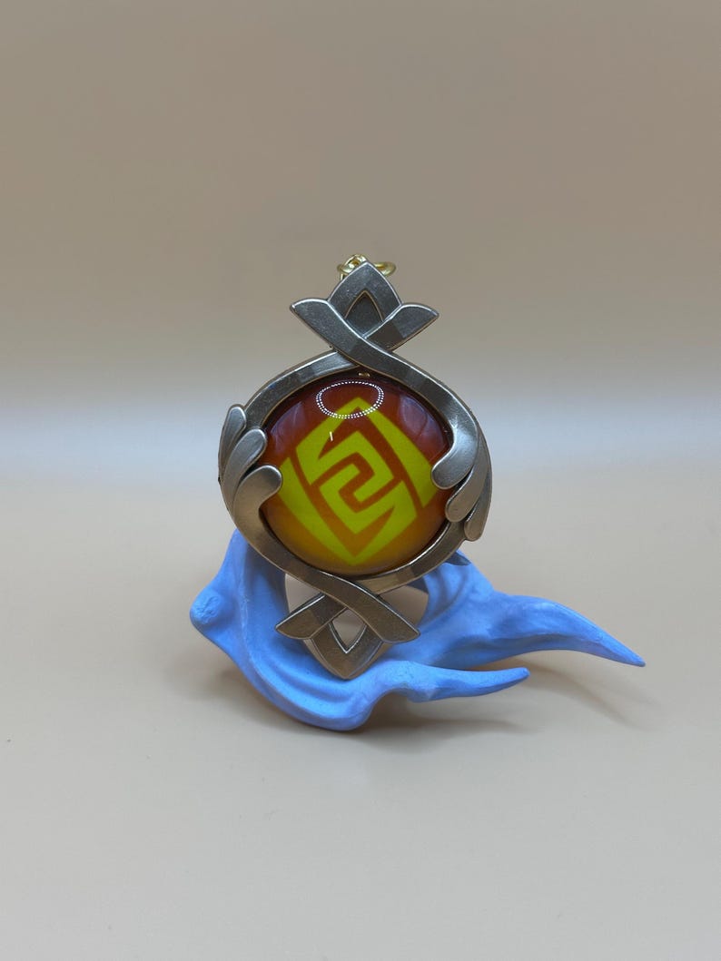 May include: A pendant with a circular, orange and yellow design, framed by silver-colored metal with decorative accents. The pendant rests on a light blue, stylized base. The design features a geometric pattern.
