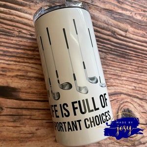May include: A white insulated tumbler featuring golf clubs and the text "LIFE IS FULL OF IMPORTANT CHOICES." The design includes black and gray golf club illustrations. The tumbler has a silver lid.