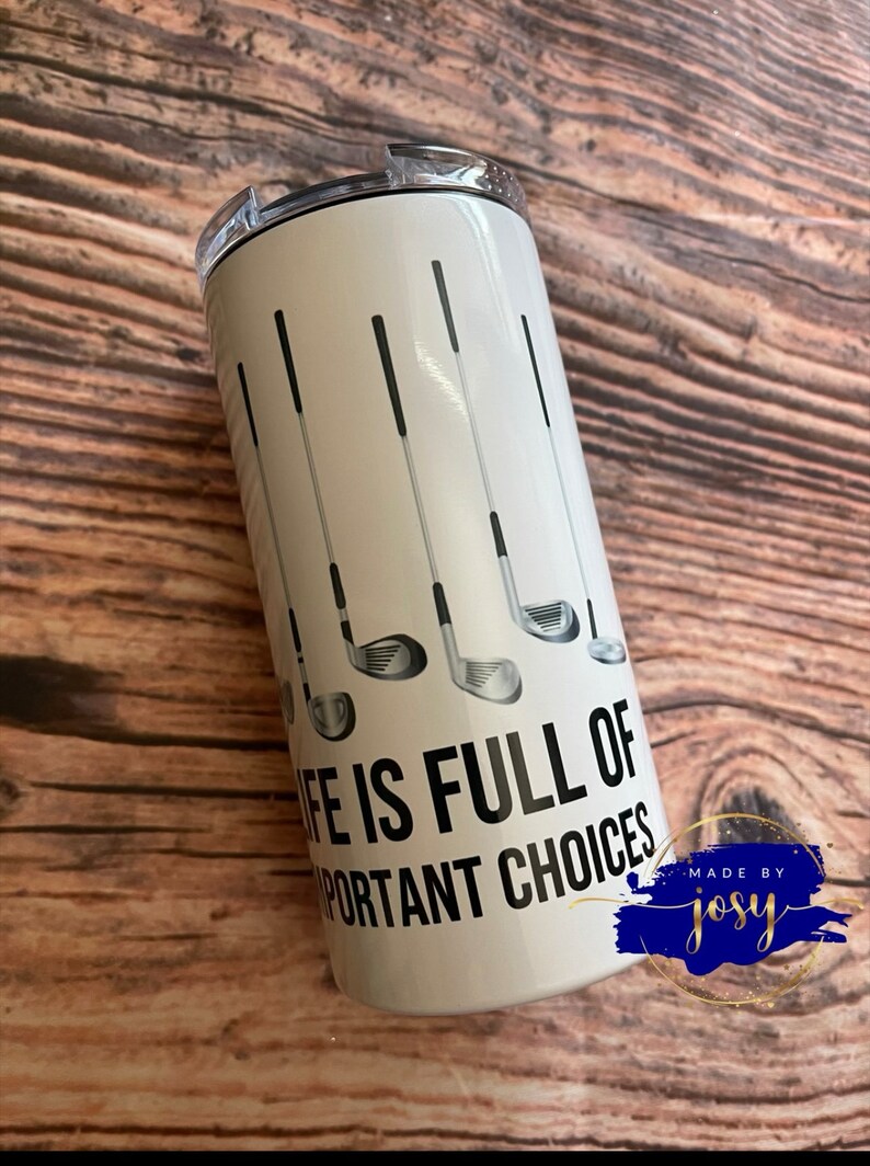 May include: A white insulated tumbler featuring golf clubs and the text "LIFE IS FULL OF IMPORTANT CHOICES." The design includes black and gray golf club illustrations. The tumbler has a silver lid.