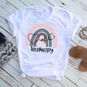 以下が含まれることがあります： 虹のデザインと聴診器が付いたハートが描かれた白いTシャツ。虹の下に「Telemetry」という文字が印刷されています。
