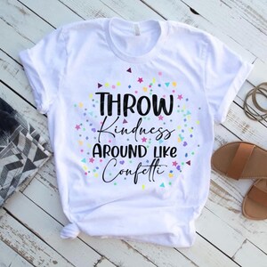 Könnte beinhalten: Weißes T-Shirt mit dem Text "Throw Kindness Around Like Confetti" in schwarzer Schrift. Der Text ist von buntem Konfetti umgeben.