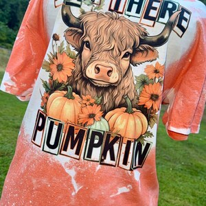 Può includere: Una t-shirt arancione candeggiata con una grafica di una mucca delle Highland circondata da fiori arancioni e zucche. Il testo "HAY THERE" è stampato in nero sopra la mucca e la parola "PUMPKIN" è stampata in nero sotto la mucca.