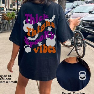 May include: Black t-shirt with a graphic print of a ghost, bats, and the text "Thick Thighs Spooky Vibes". The graphic is in purple, orange, and white. The shirt is oversized and has a weightlifting theme.