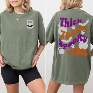 May include: A green oversized t-shirt with a black kettlebell graphic on the front and the text "Thick Thighs Spooky Vibes" with a purple and orange color scheme on the back.