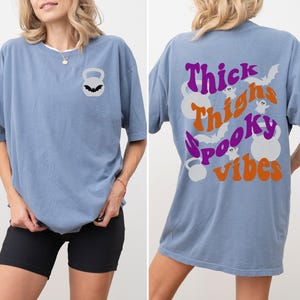 May include: A light blue oversized t-shirt with a graphic print on the back that says "Thick Thighs Spooky Vibes" with bats and ghosts. The front of the shirt has a small graphic of a kettlebell with a bat on it.
