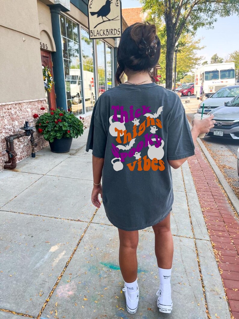 May include: A dark gray t-shirt with a graphic print that says "Thick Thighs Spooky Vibes" with a ghost and bats. The graphic also includes a purple and orange kettlebell.