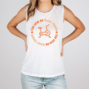 May include: White sleeveless shirt with orange text and a stationary bike graphic. The text around the bike reads "You spin me right round". The shirt is a casual style, suitable for workouts or everyday wear.