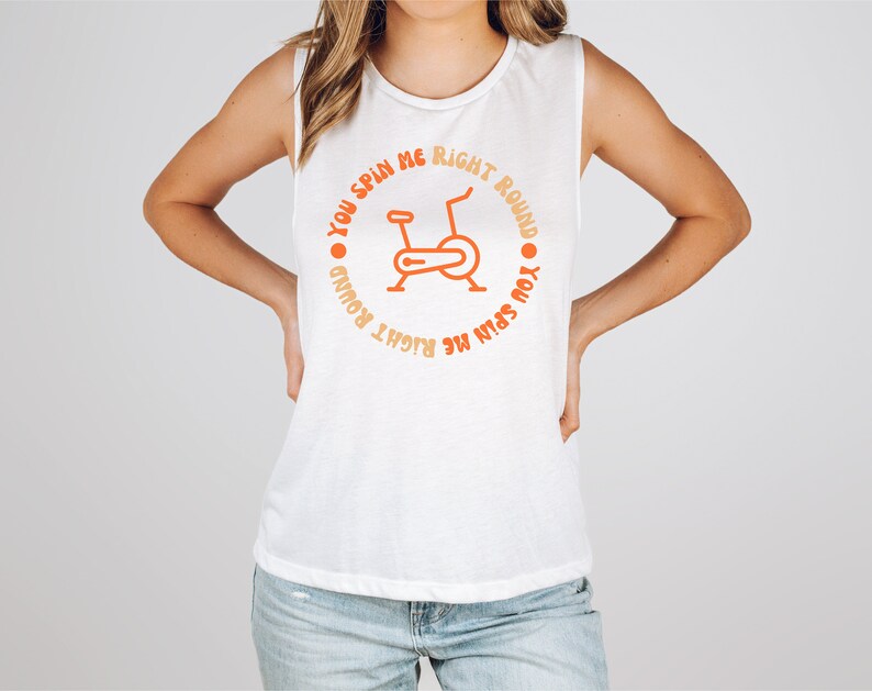 May include: White sleeveless shirt with orange text and a stationary bike graphic. The text around the bike reads "You spin me right round". The shirt is a casual style, suitable for workouts or everyday wear.