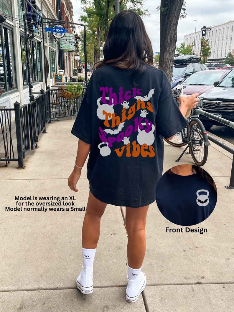 May include: Black t-shirt with a graphic print of a ghost, bats, and the text "Thick Thighs Spooky Vibes". The graphic is in purple, orange, and white. The shirt is oversized and has a weightlifting theme.
