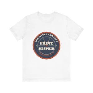 May include: A white short-sleeve t-shirt featuring a circular graphic on the chest. The design has a dark blue center with the text "PAINT & DESPAIR" in white, surrounded by a red scalloped border and the words "MINIATURE PAINTERS" and "LOCAL BRANCH NO. 20".