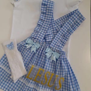 May include: A blue and white gingham dress with a white collared shirt, white socks, and blue bows. The dress has a white lace trim and the word "JESUS" written in gold letters on the front.