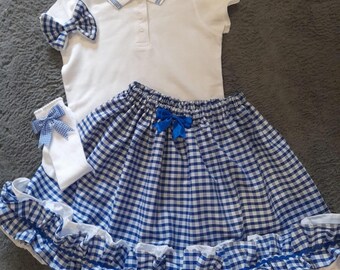 May include: A white polo shirt with blue trim, a blue and white gingham skirt with a blue bow, a matching bow, and white socks with a blue gingham bow. The skirt has a ruffled hem.