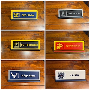 Puede incluir: Seis placas de escritorio con insignias y rangos militares. Las placas son de varios colores, incluyendo azul, amarillo, rojo y blanco, con texto negro. Cada placa muestra un nombre y un rango militar.