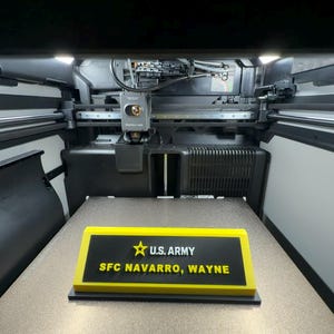 Op de afbeelding: Een rechthoekig naambordje met een zwart midden en een felgele rand. De tekst luidt U.S. ARMY met een geel sterpictogram, gevolgd door de naam SFC NAVARRO, WAYNE in gele letters. Het object bevindt zich in een 3D-printer.