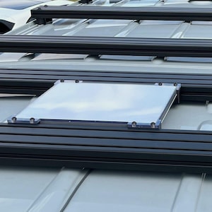 Könnte beinhalten: Ein Fahrzeug-Dachträgersystem mit schwarzen Querträgern und einer transparenten, rechteckigen Platte. Die Platte ist mit silbernen Beschlägen befestigt. Der Träger ist auf einem hellfarbigen Fahrzeugdach montiert.