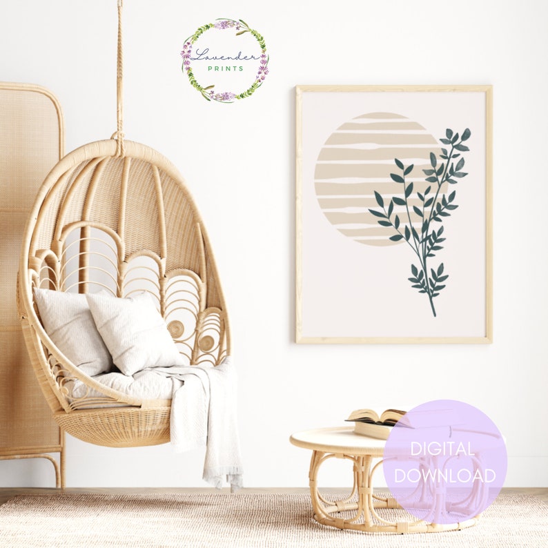 May include: A digital download of a minimalist art print featuring a green leaf branch against a beige background with a circle and stripes. The print is framed and hanging on a white wall above a wicker coffee table.